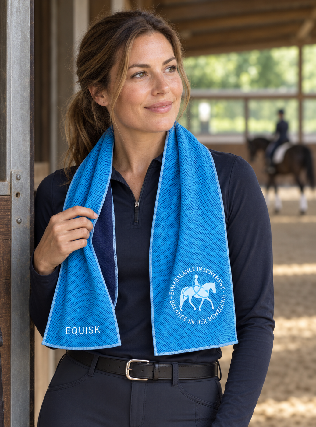 A woman in equestrian apparel stands in a stable doorway wearing a bright blue towel draped around her neck. The towel has white branding text reading 'EQUISK' on one side and a white horse-and-rider logo with circular text on the other.