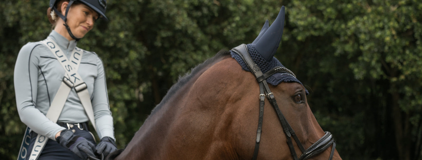 Woman in equestrian gear standing next to a horse with a blurred green background
